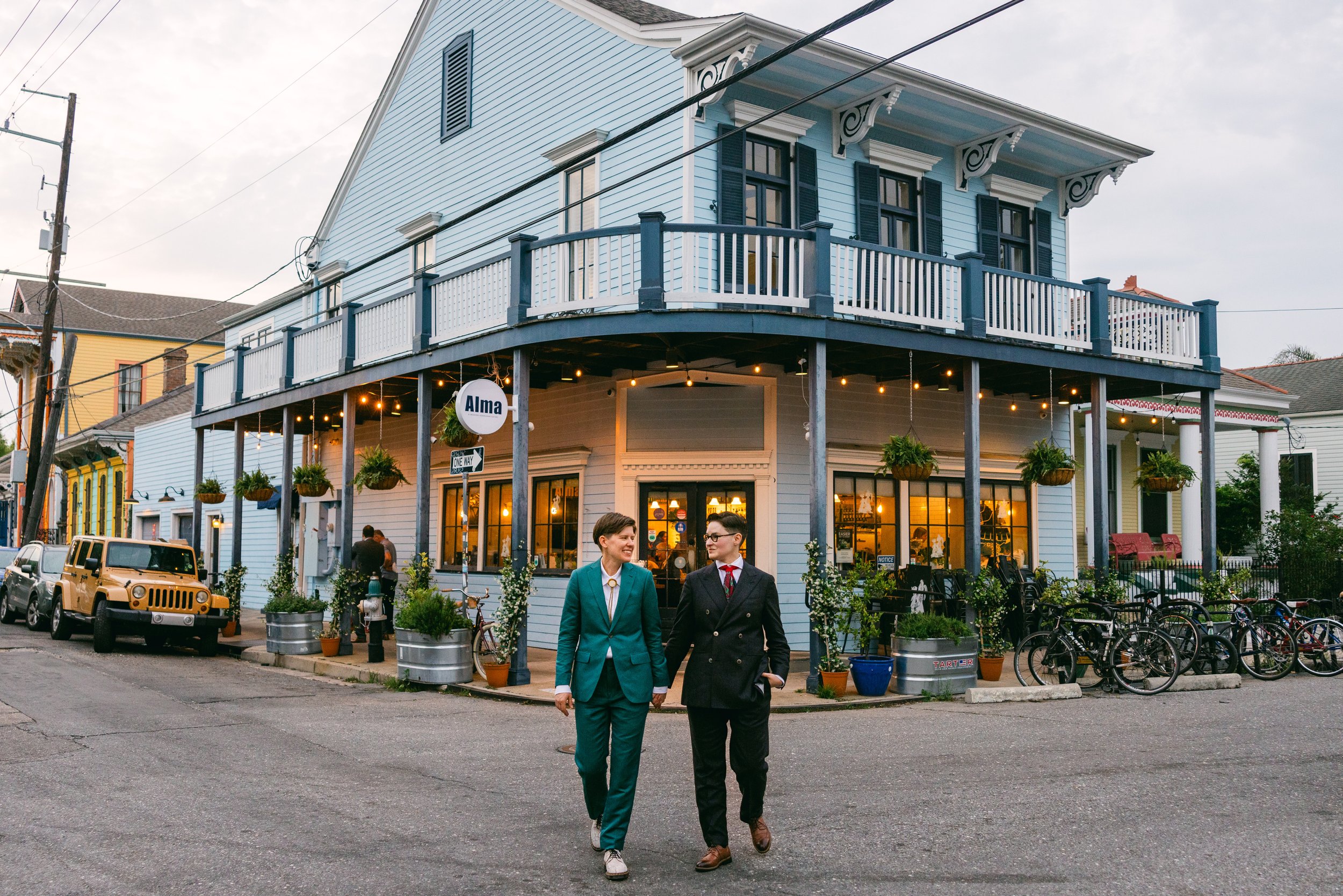 Two people dressed in formal attire, a man and a woman, walking and talking on a city street in front of a light blue, two-story building with outdoor balcony and hanging plants, with parked cars and bicycles nearby.