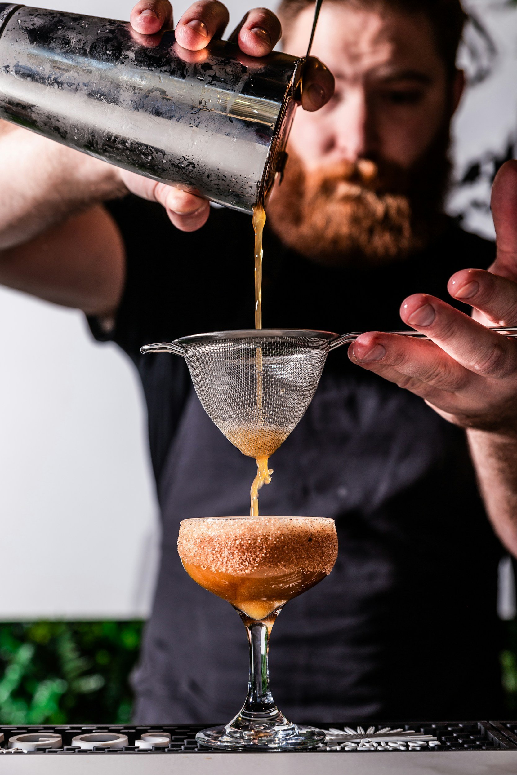 A bartender pouring brown liquid through a fine mesh strainer into a cocktail glass layered with sugar, on a bar.