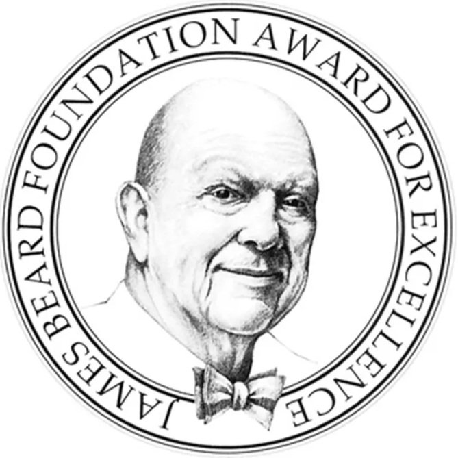 Black and white circular logo featuring a portrait of an elderly man with a bald head, wearing a bow tie. The text around the logo reads 'IMME'S BEARD FOUNDATION AWARD FOR EXCELLENCE'.