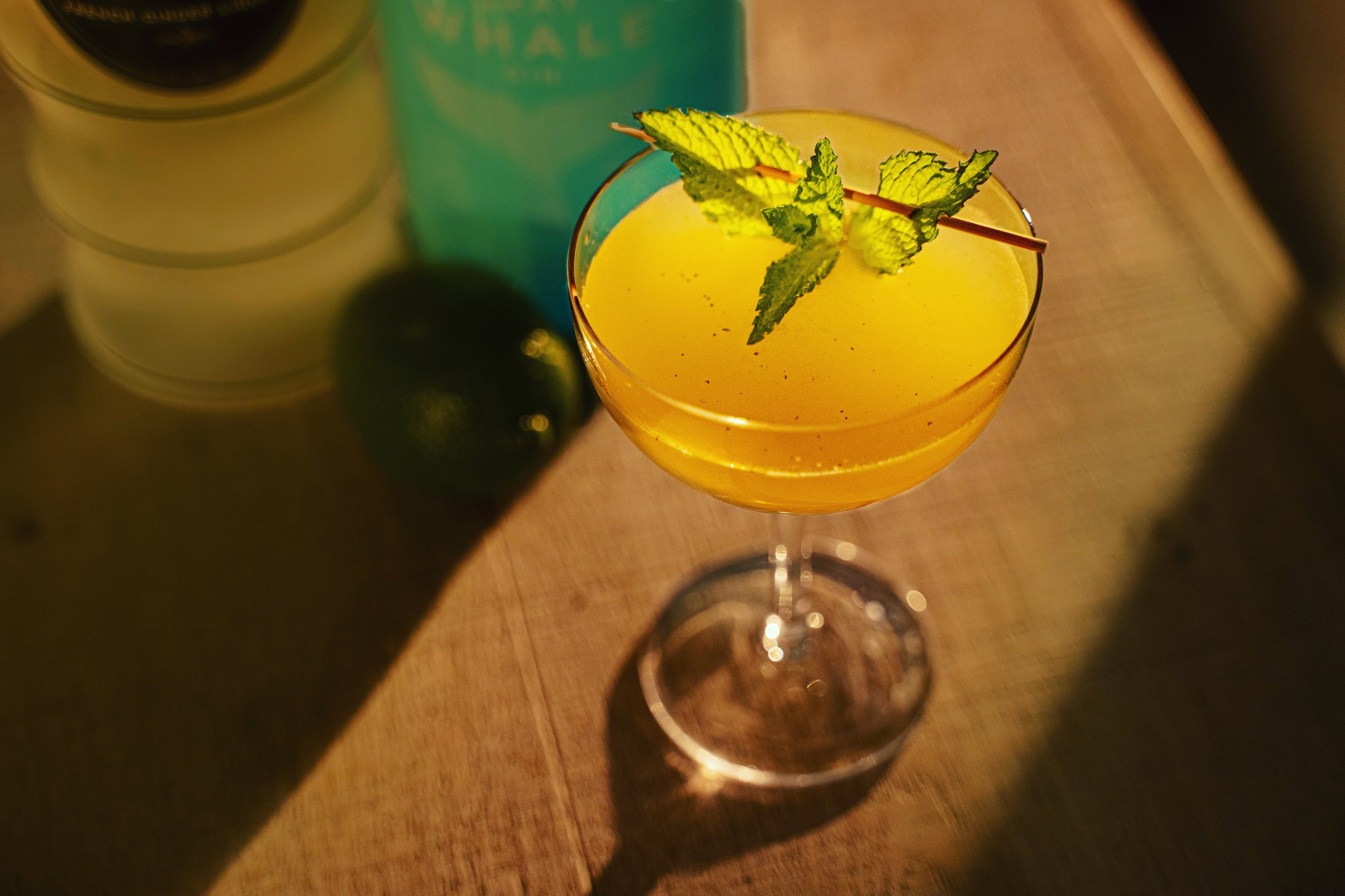 A cocktail in a wine glass garnished with a sprig of fresh mint, placed on a wooden surface with a can and a lime in the background.