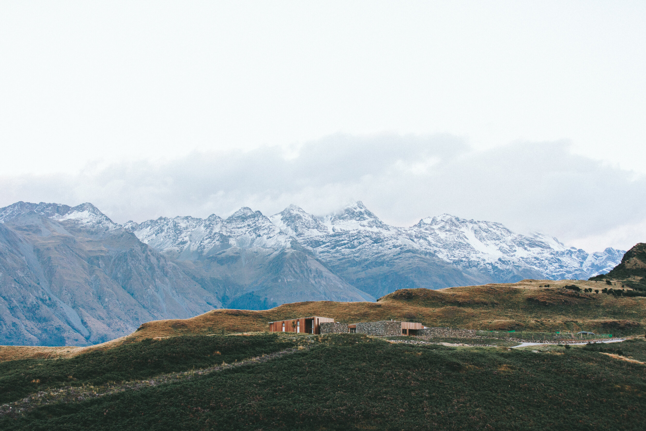 A mountain landscape with snow-capped peaks in the background, rolling green hills in the foreground, and a modern building situated on the hills.
