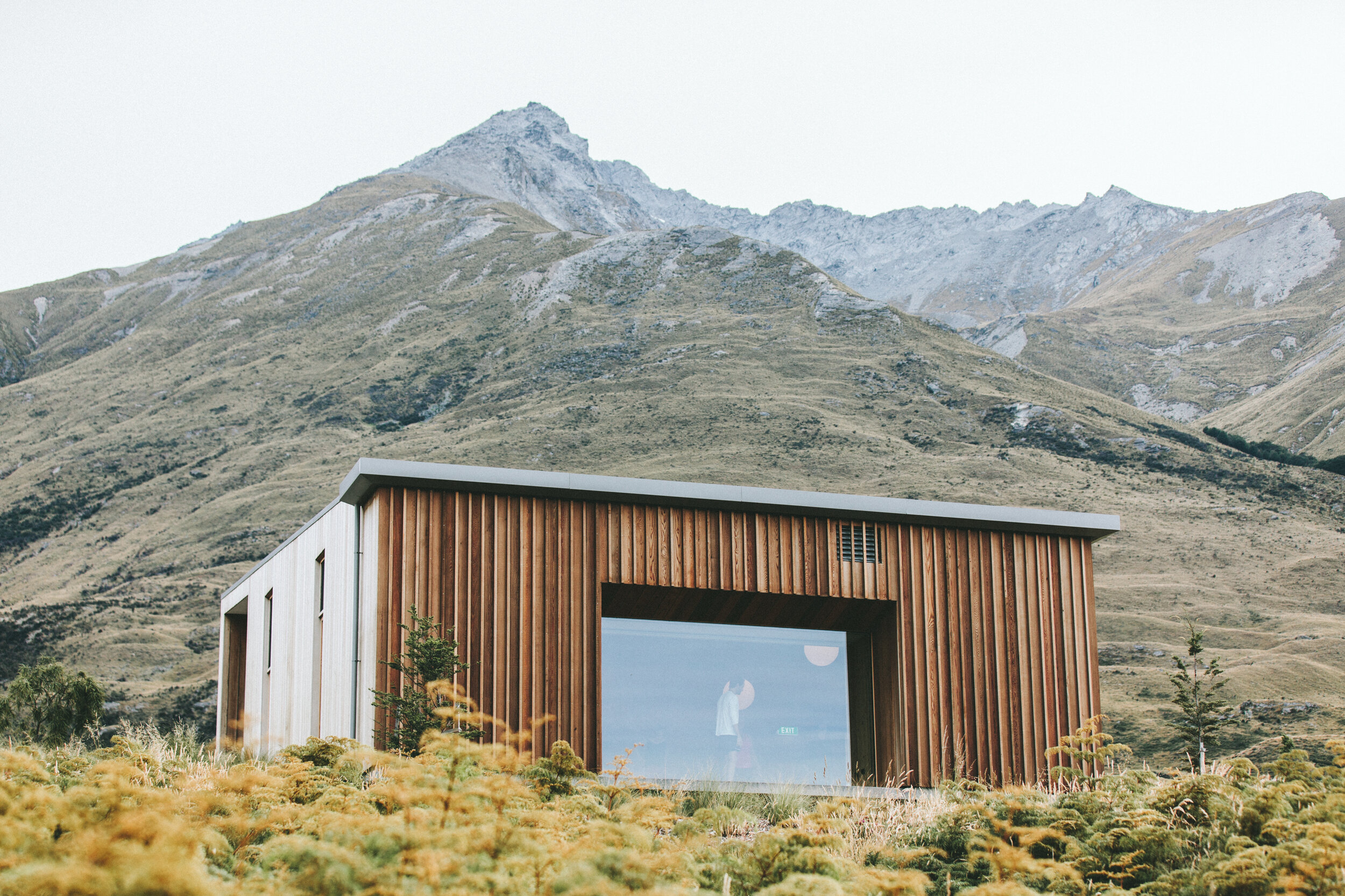 Modern wooden building with large glass window set against mountainous landscape with rolling hills and rocky peaks.