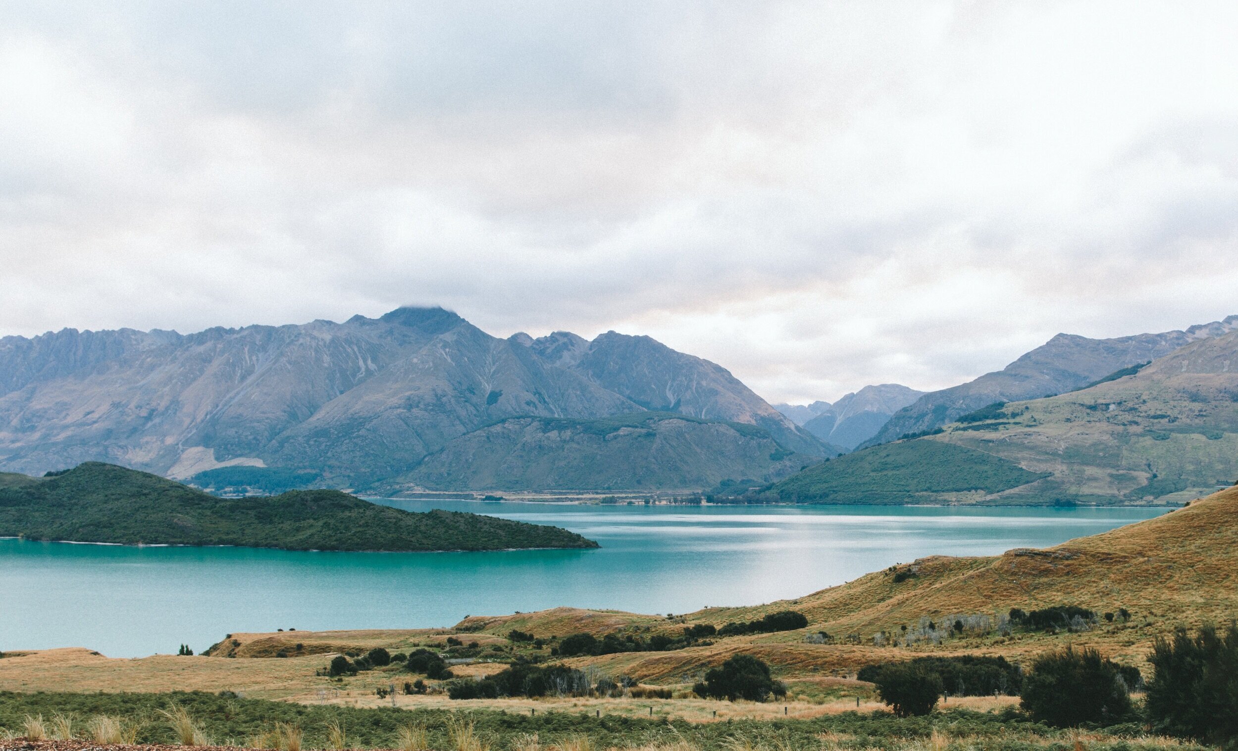 Scenic landscape of mountains, green hills, and a lake under cloudy sky.